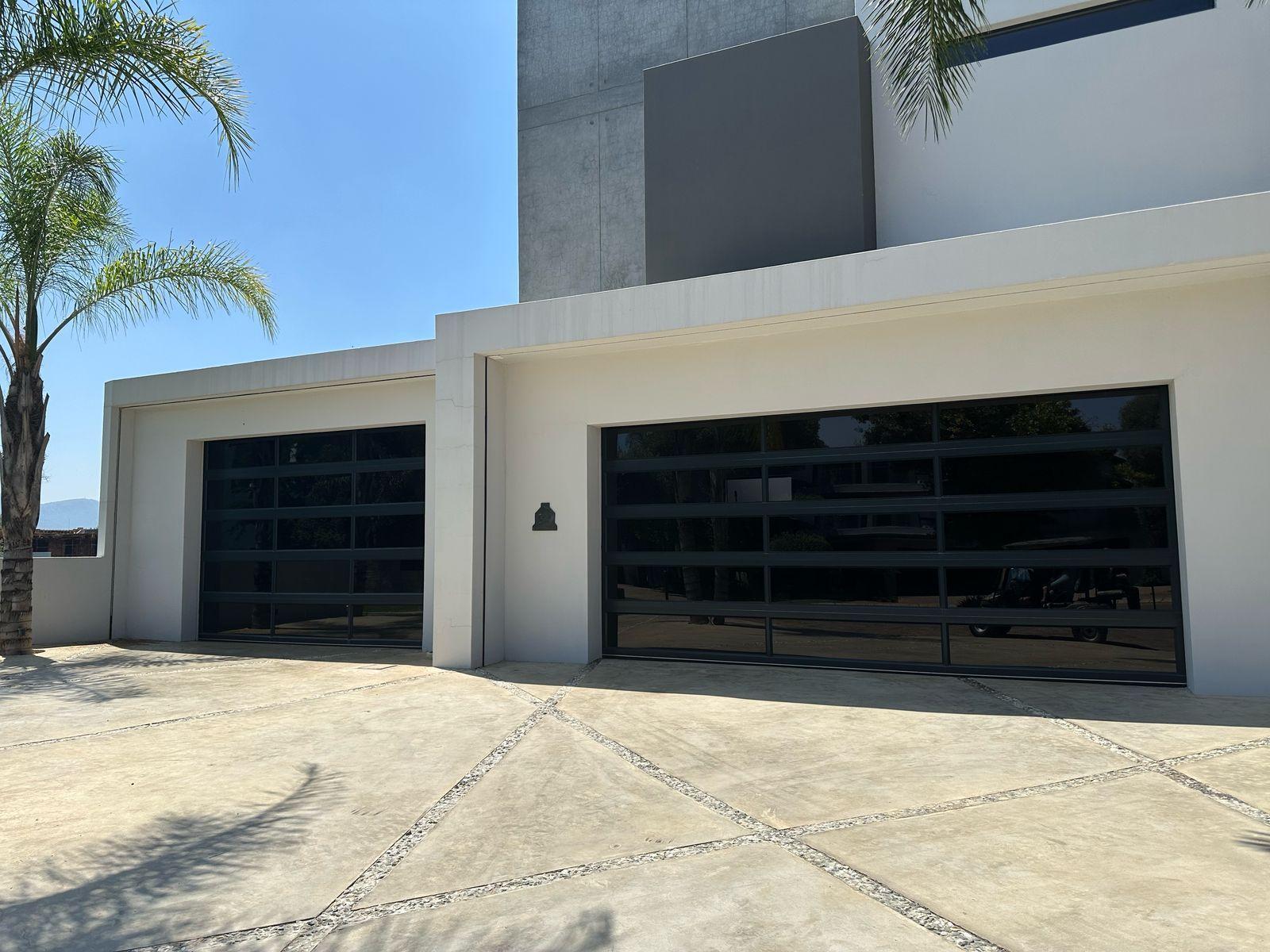 Modern house exterior with two large garage doors, palm tree and concrete driveway under bright blue sky.